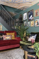 Salon vert ouvert avec des canapés en velours rouge et vert devant un escalier ouvert et un mur d'exposition d'art de style salon et un plafond à motifs en mosaïque