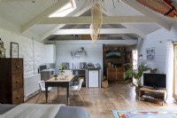 Grange convertie, salon ouvert avec cuisine simple et table de ferme. Poutres en bois et décoration rustique.